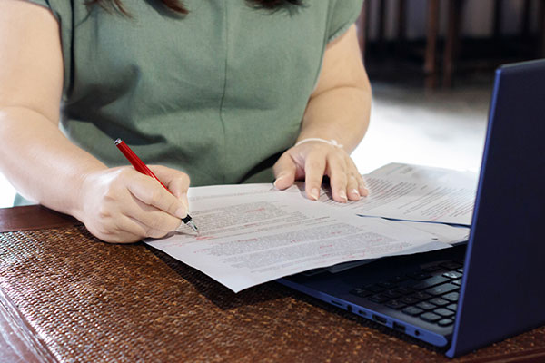 Persons hand holding pen marking on proofreading paper with laptop on desk in office