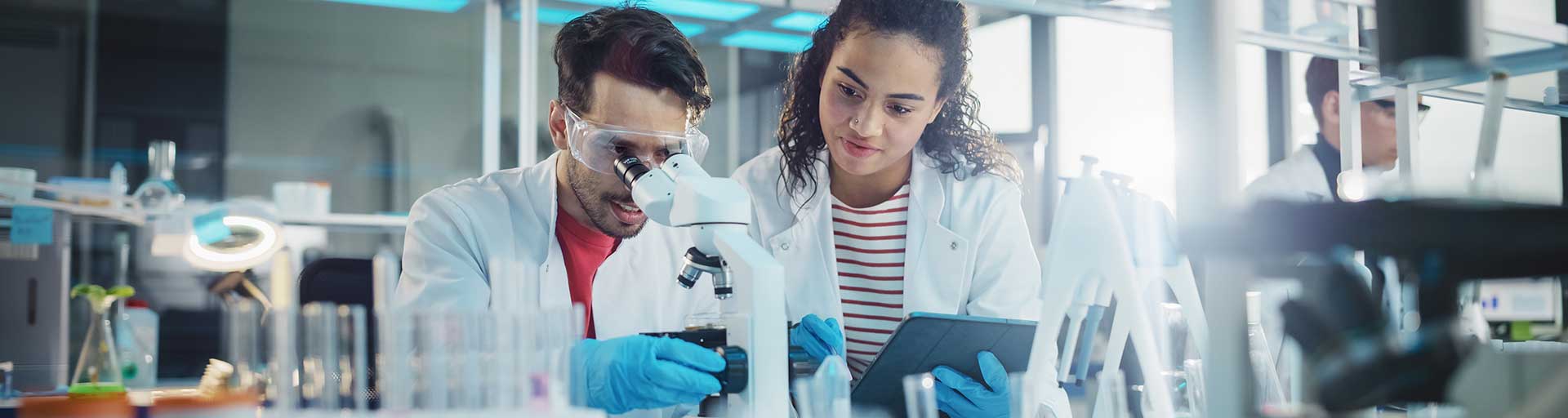 students in lab coats working with a microscope and tablet in a lab